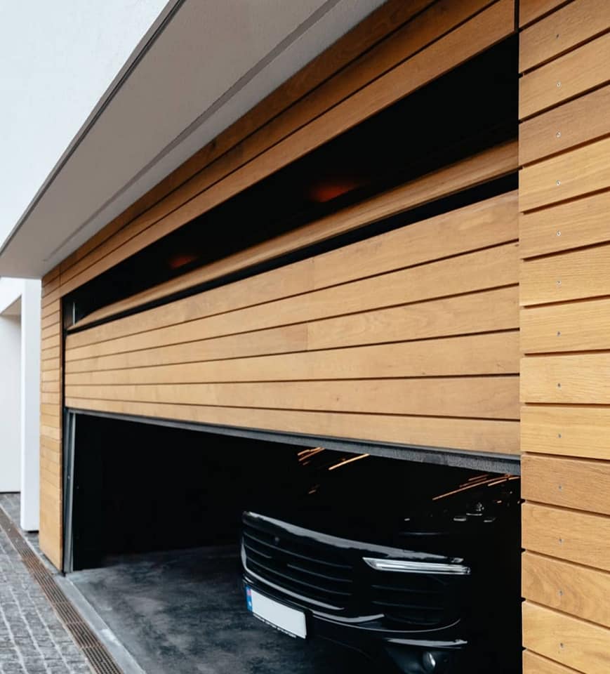 Modernes Garagentor aus horizontalen Holzlatten, halb geöffnet, mit dahinter geparktem schwarzen Fahrzeug.